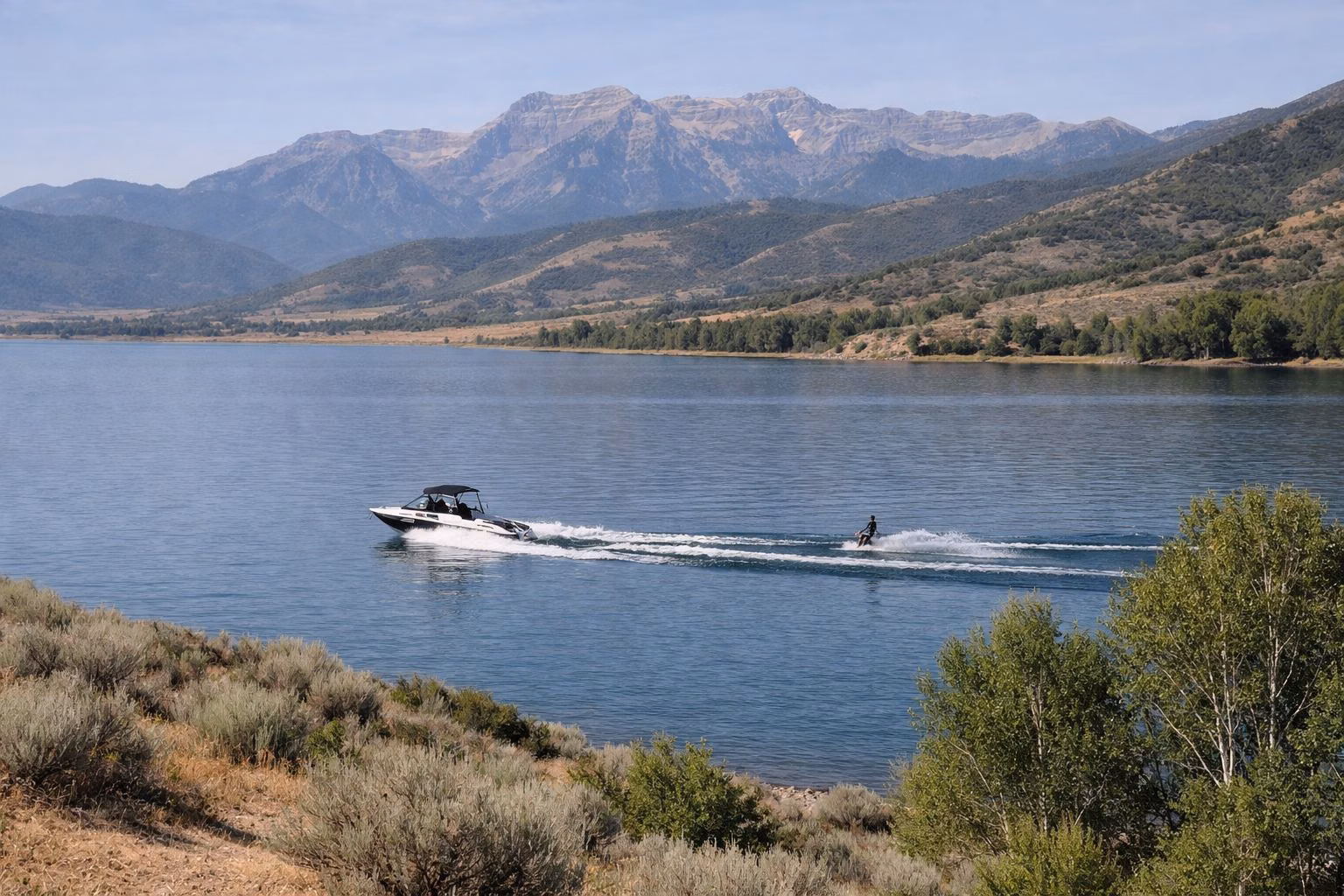 A ski boat towing a wakeboarder across a calm mountain reservoir