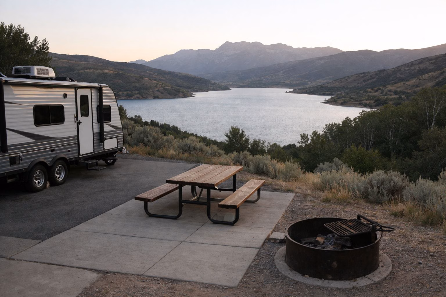A hillside RV campsite with a picnic table and fire ring overlooking a blue mountain reservoir
