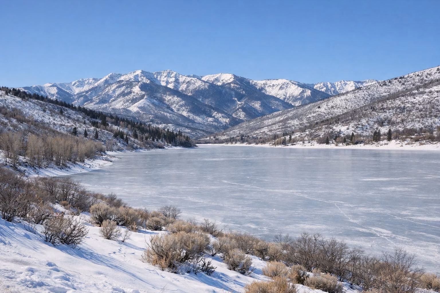 A frozen snow-covered mountain reservoir with snowy hills and mountains behind