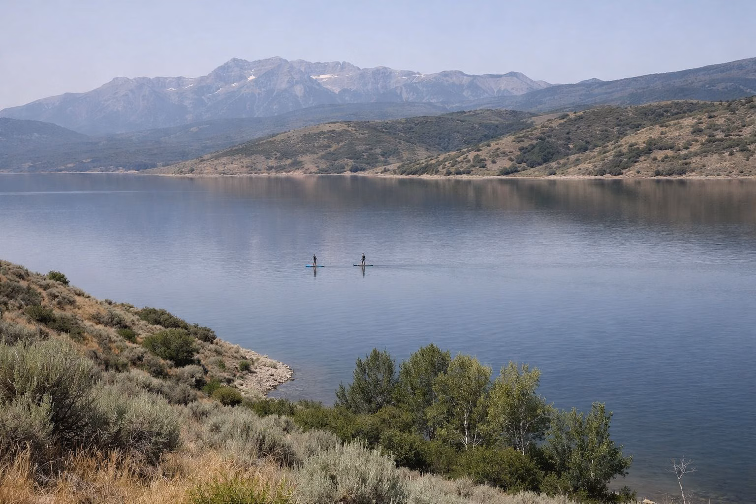 Two distant stand-up paddleboarders on a glassy mountain reservoir