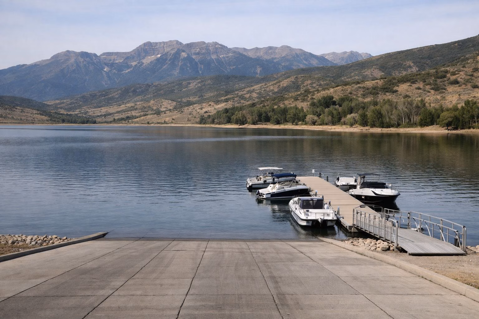 A concrete boat ramp and floating dock with moored motorboats on a calm mountain reservoir
