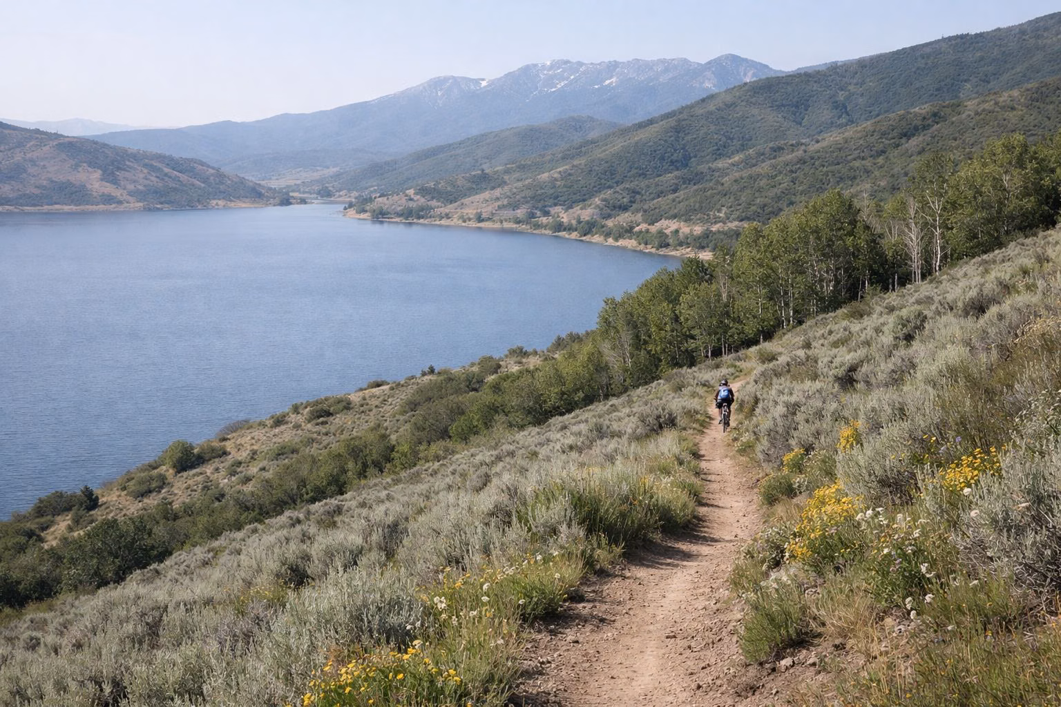 A dirt singletrack trail across a sagebrush hillside above a blue reservoir, a mountain biker in the distance