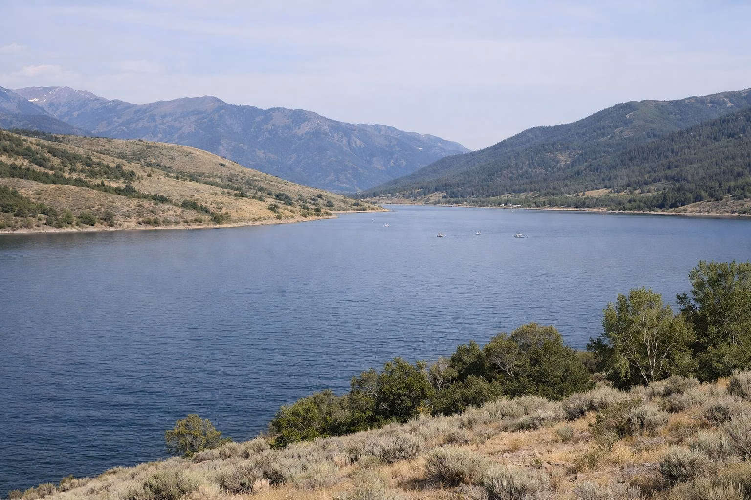 A wide mountain reservoir with sagebrush hills and forested peaks behind, small boats far out on the water