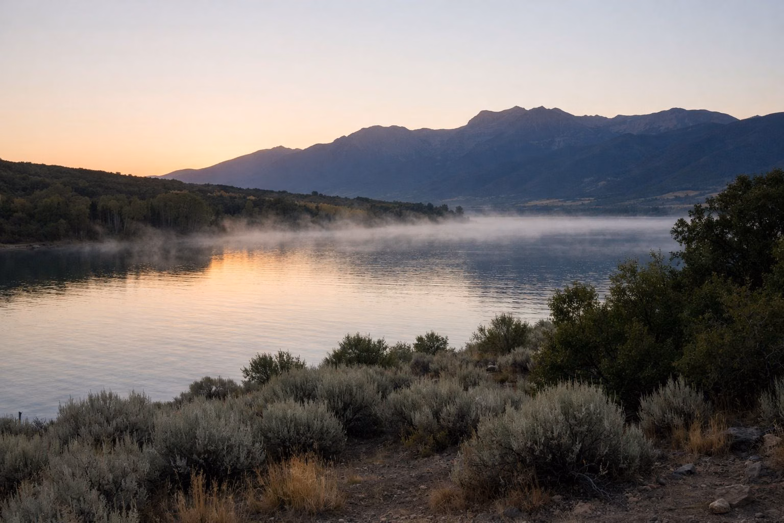 Sunrise over a calm mountain reservoir with mist on the water and a sagebrush shoreline