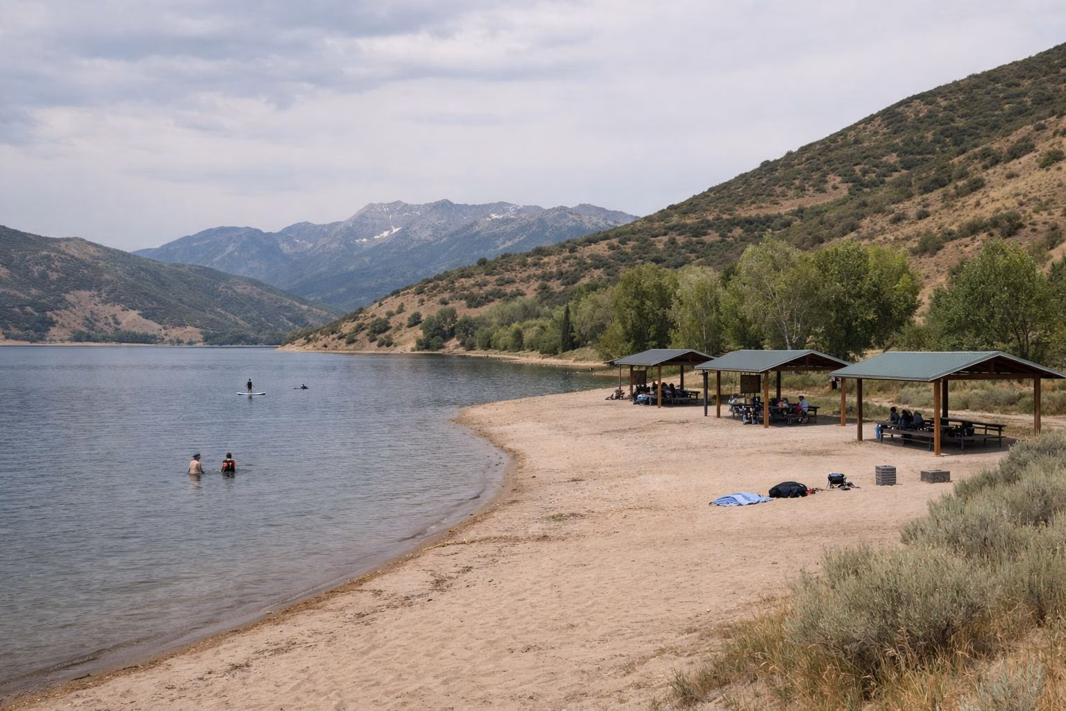 A small sandy beach on a mountain reservoir with covered picnic shelters and a few distant swimmers