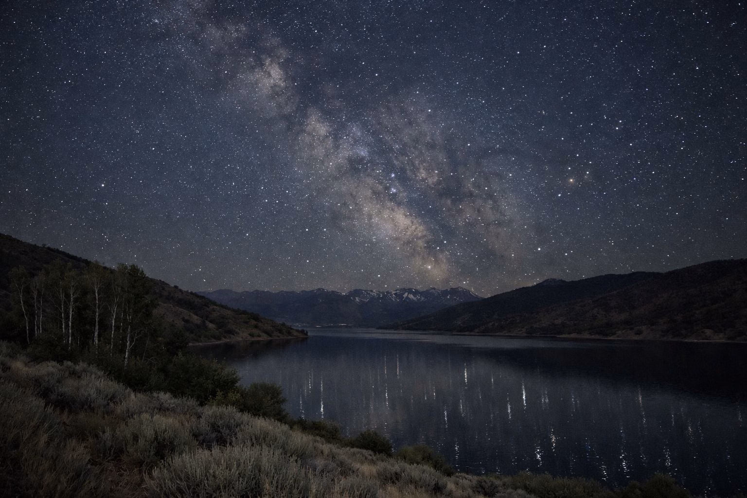 A starry night sky and the Milky Way over a calm mountain reservoir