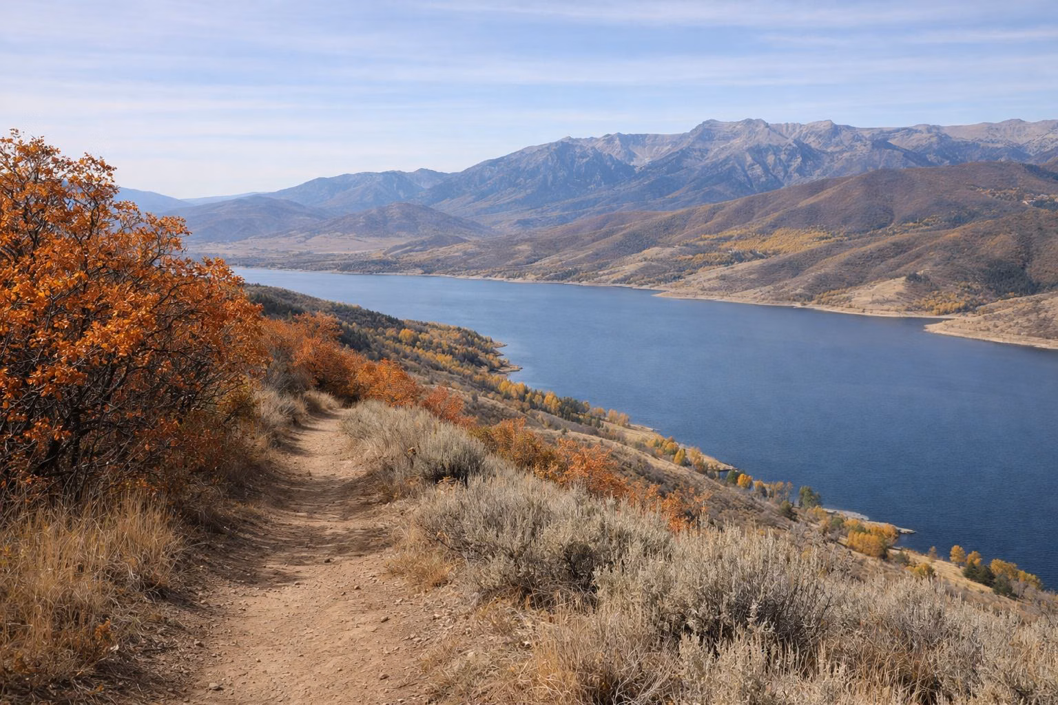 A dirt trail along a ridge of red-orange scrub oak above a large reservoir